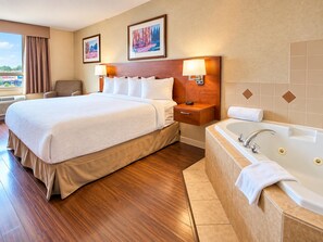 Habitación de hotel con una cama grande, una mesita de noche con lámpara, un baño con bañera y dos cuadros en la pared.