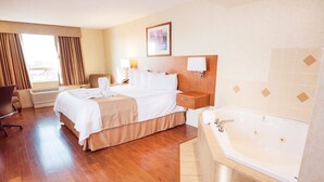 A hotel room with a large bed, a desk, a chair, a desk lamp, a bedside table, a wall-mounted lamp, and a bathtub. - Rosslyn Inn and Suites (Edmonton)