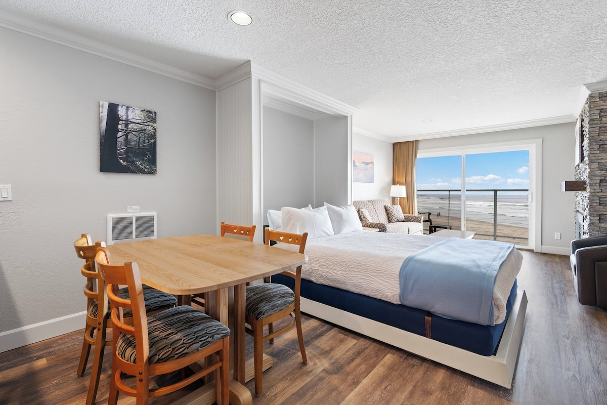 A hotel room with a large bed, a dining table with chairs, a view of the ocean, and a fireplace.