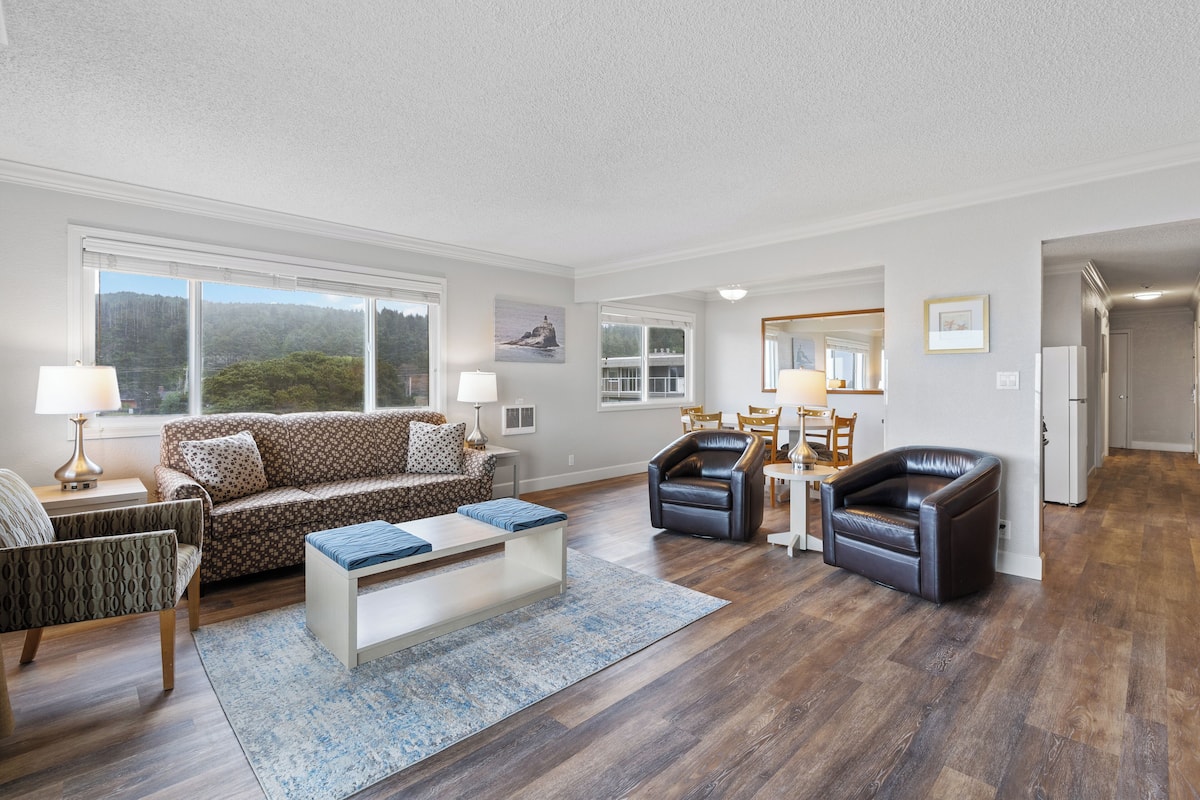 A spacious living room with a sofa, coffee table, and chairs. There are large windows with a view of trees and hills.