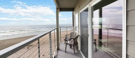 A balcony with a view of the beach, a railing, and two chairs.