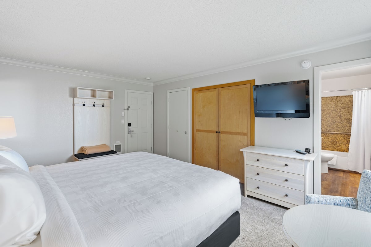 A hotel room with a large bed, a TV mounted on the wall, a wooden wardrobe, a white dresser, and a chair.