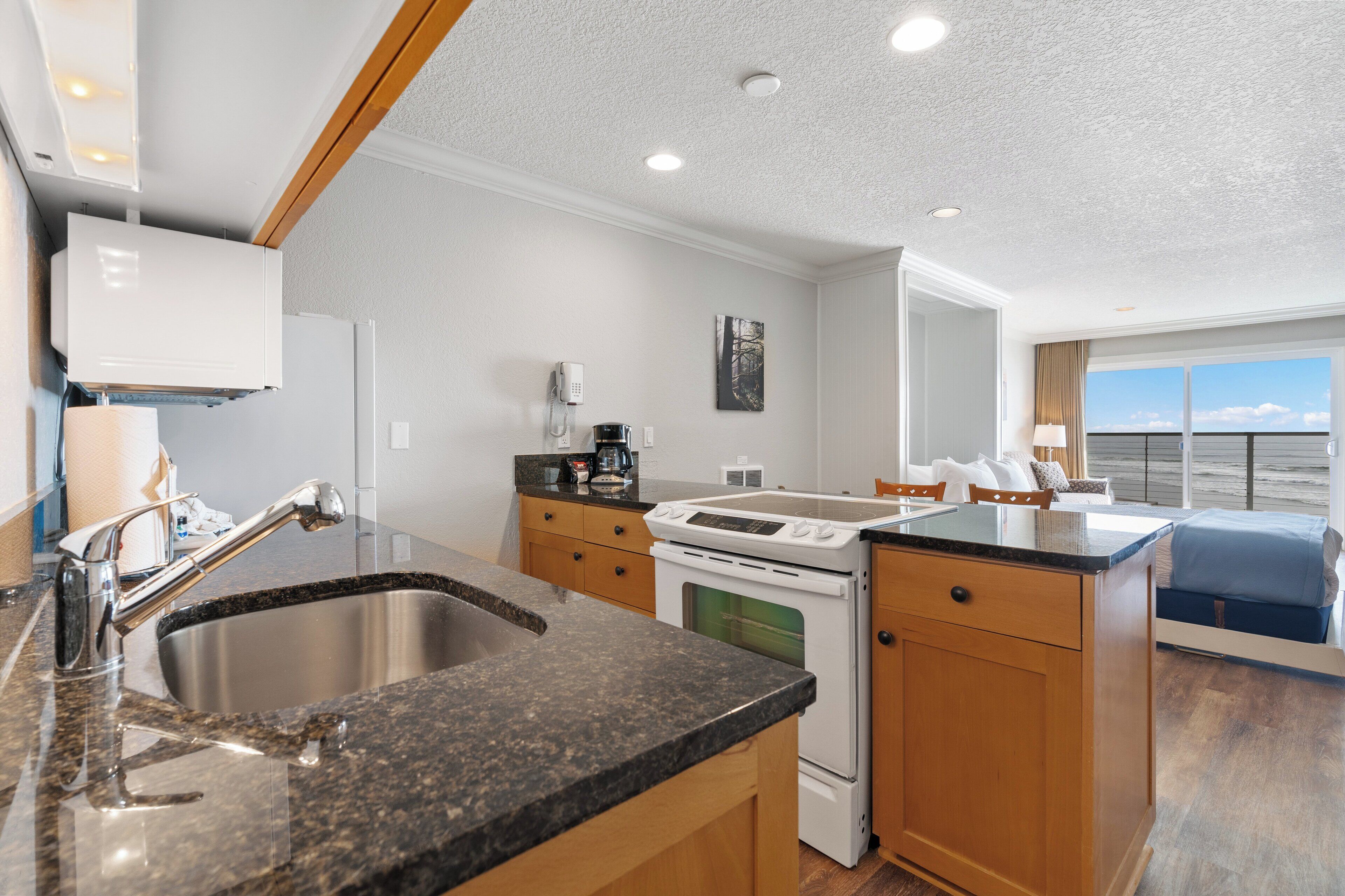 A modern kitchen with granite countertops, stainless steel sink, and white appliances. There is a dining area with a blue chair and a view of the outdoors.