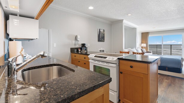 A modern kitchen with granite countertops, stainless steel sink, and white appliances. There is a dining area with a blue chair and a view of the outdoors.