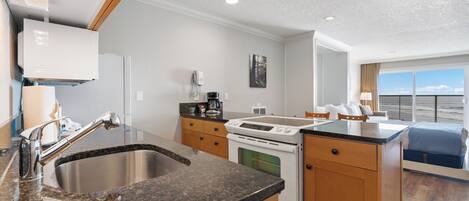 A modern kitchen with granite countertops, stainless steel sink, and white appliances. There is a dining area with a blue chair and a view of the outdoors.
