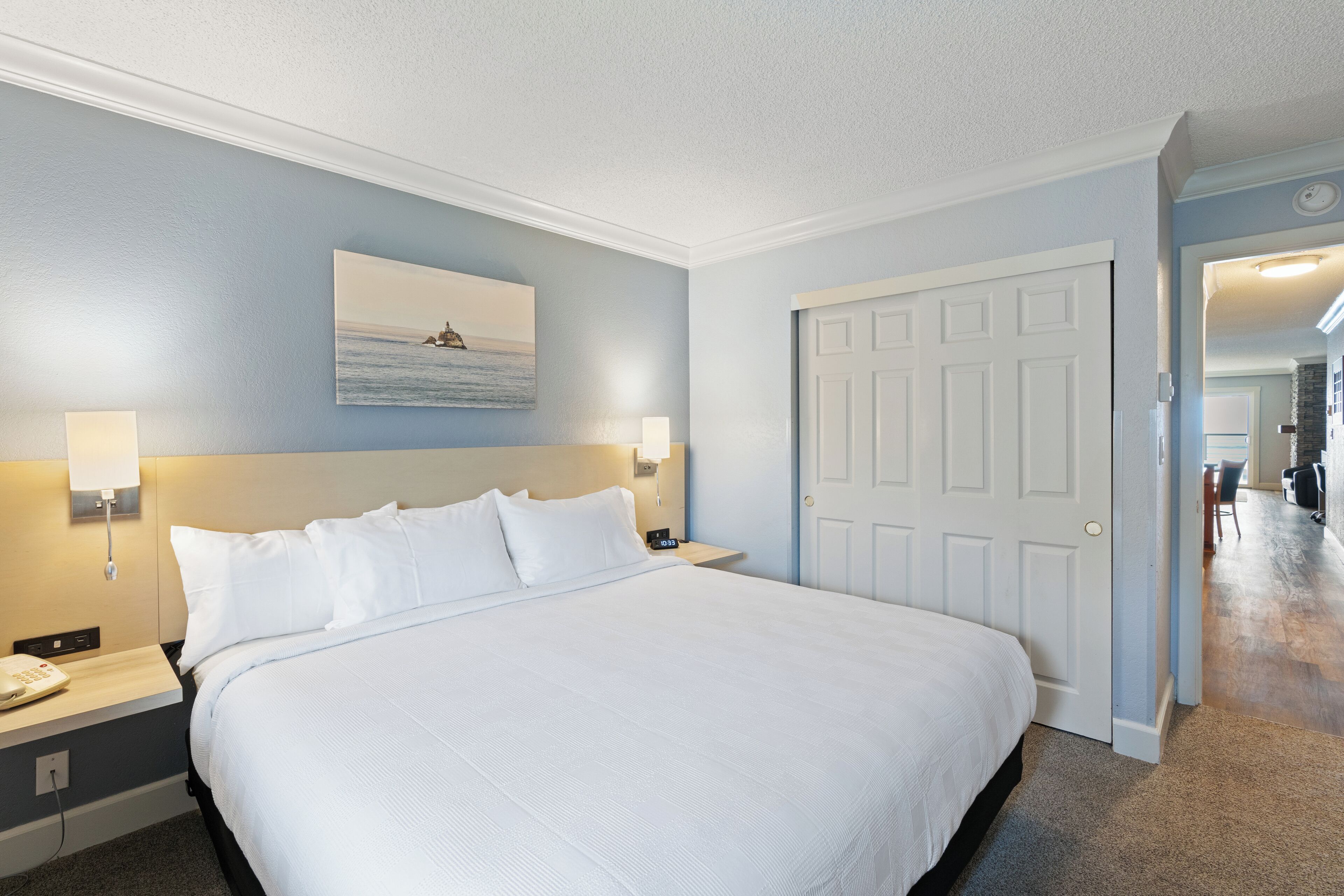 A hotel room with a large bed, a nightstand with a phone, a wall-mounted lamp, and a door leading to another room.