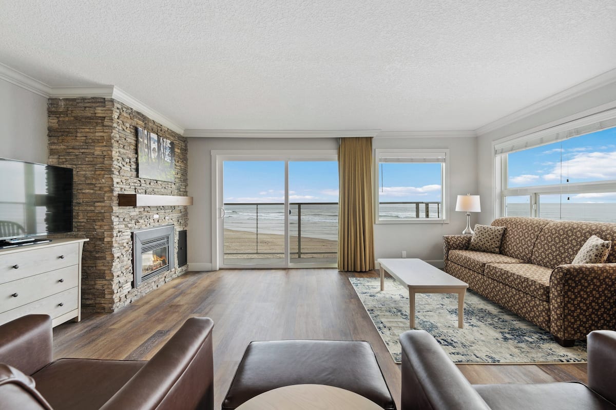 A living room with a stone fireplace, a television, a sofa, and a view of the beach through large windows.