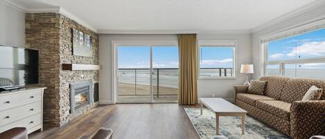A living room with a stone fireplace, a television, a sofa, and a view of the beach through large windows.