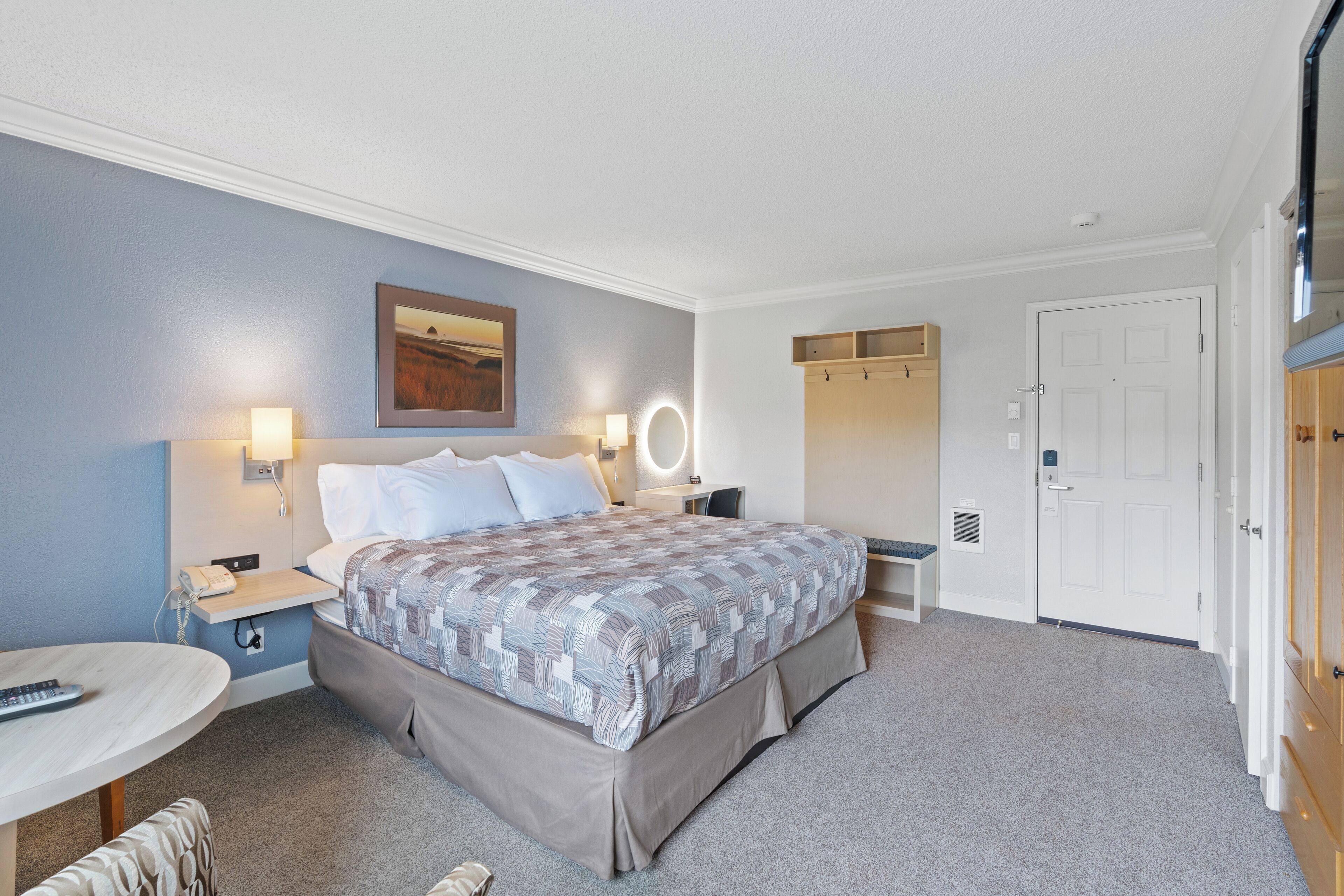 A hotel room with a bed, bedside tables, a desk, and a chair. There is a door and a coat rack.
