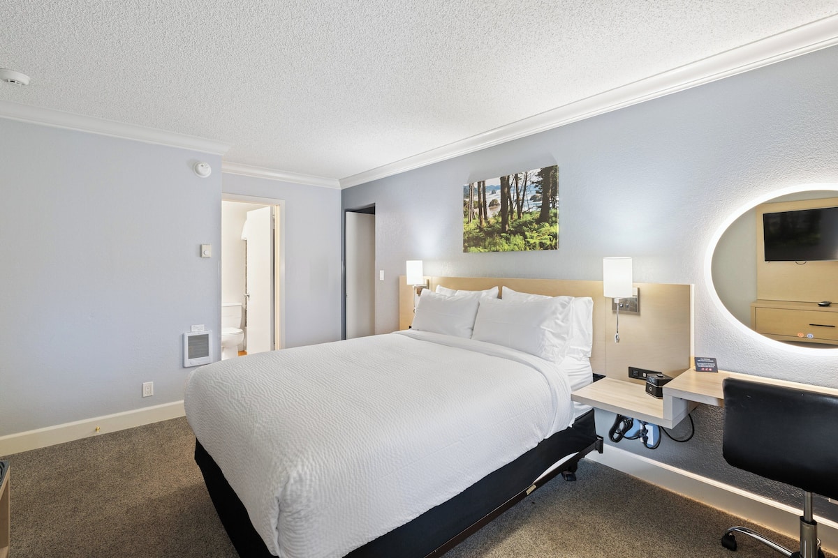 A hotel room with a large bed, bedside tables, a desk with a chair, a round mirror, and a painting on the wall.