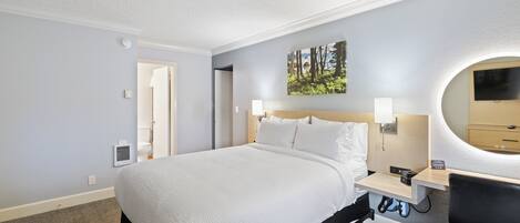 A hotel room with a large bed, bedside tables, a desk with a chair, a round mirror, and a painting on the wall.