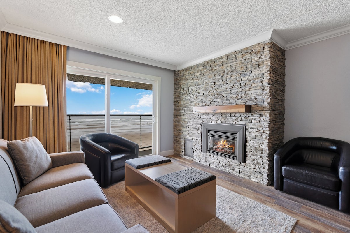 A modern living room with a stone fireplace, leather sofas, and a large window offering a view.