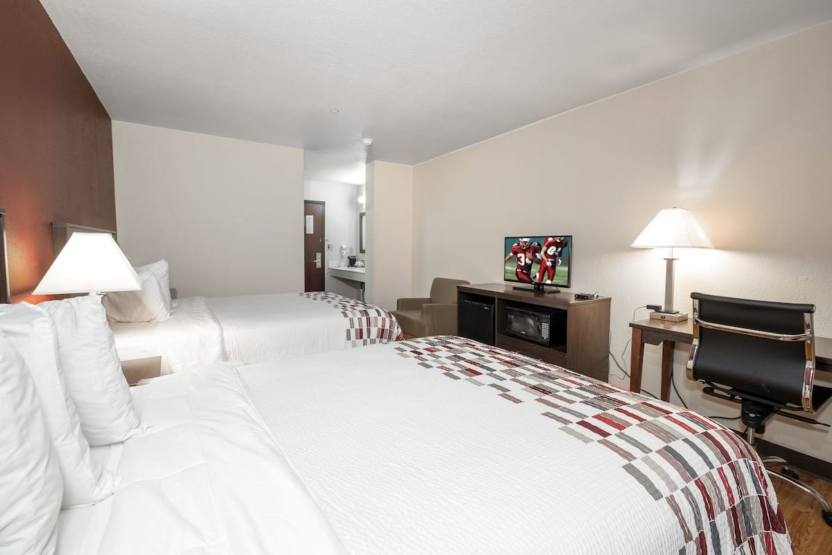 A hotel room with two beds, a desk, a chair, a TV, and a lamp.