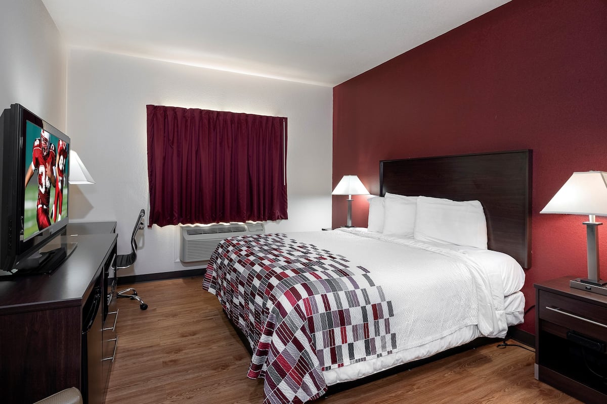 A hotel room with a bed, a TV on a stand, a desk with a lamp, and a window with curtains.
