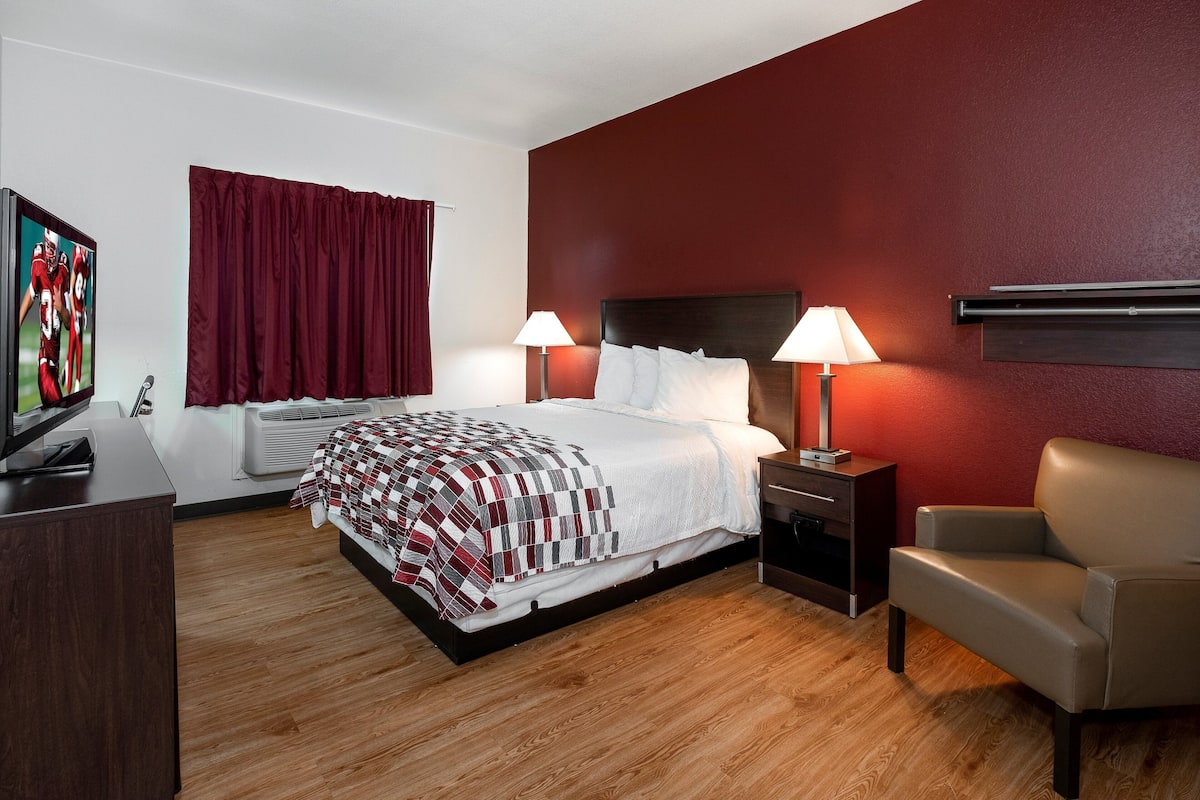 A hotel room with a large bed, two bedside tables with lamps, a TV, a chair, and a red accent wall.