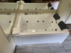 Ett stort, rektangulärt bubbelbad med flera kranar och en kontrollpanel, placerat i ett rum med kaklat golv och en stor spegel ovanför.