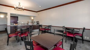 A hotel dining area with a central table surrounded by red chairs, a kitchen area with coffee machines, and a chandelier. - Red Roof Inn Hartselle (Hartselle)