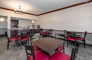 Zona de comedor de hotel con una mesa central rodeada de sillas rojas, una cocina con máquinas de café y una araña.