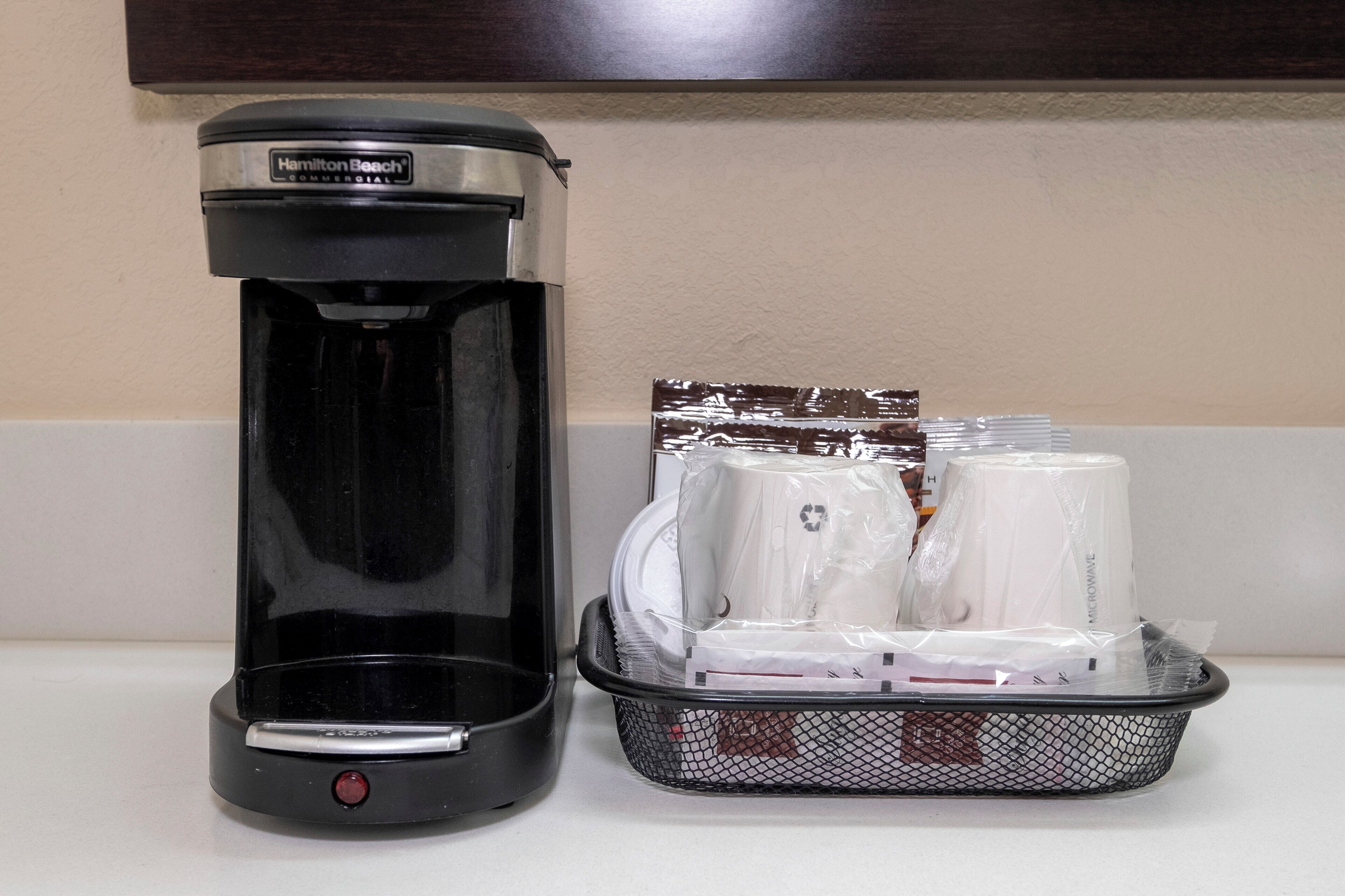 Una cafetera comercial negra y una cesta con tazas de café para microondas en una encimera.