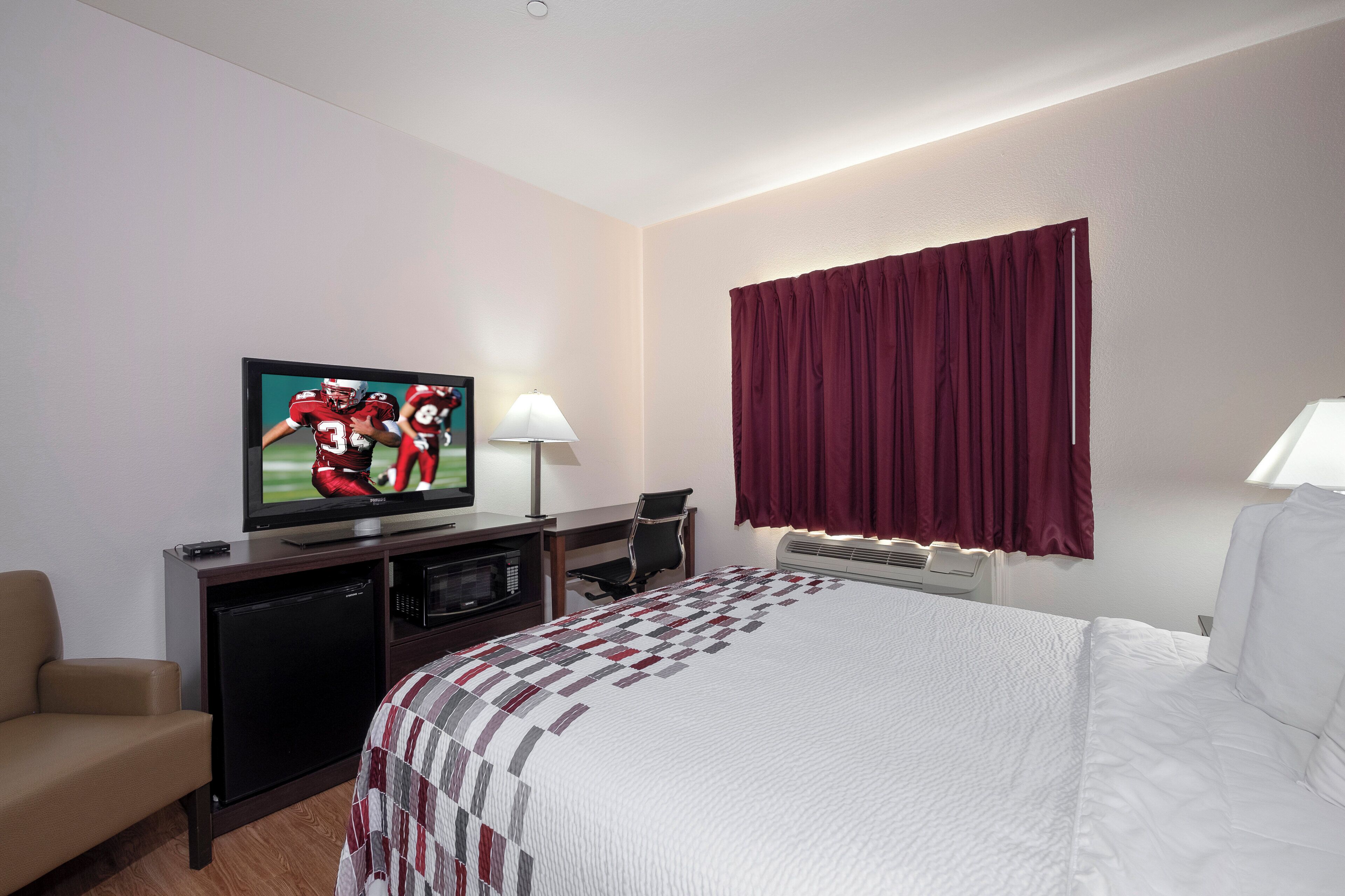 A hotel room with a television, a desk, a chair, a bed, a window with curtains, and a wall-mounted heater.
