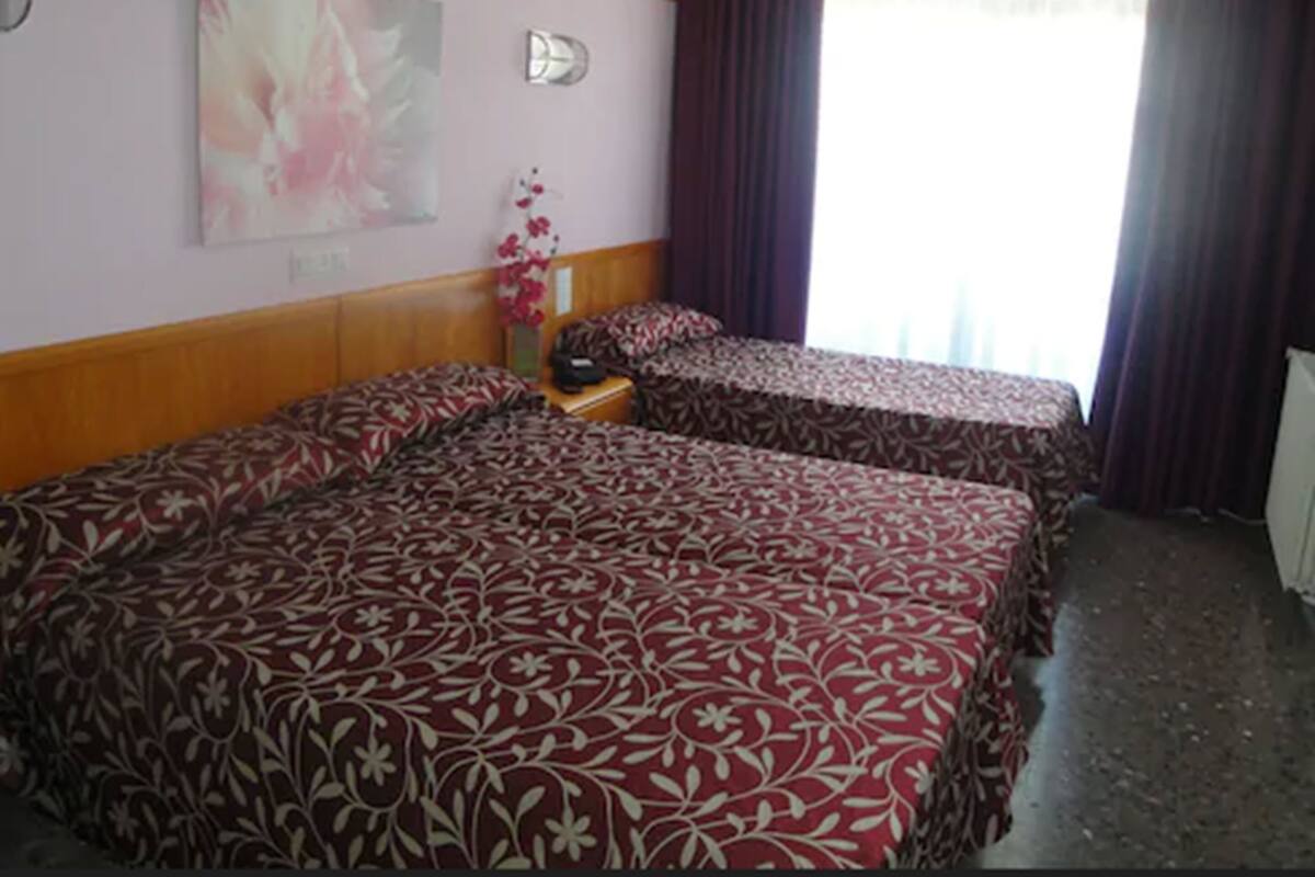 A hotel room with two beds, a wooden headboard, a painting, a vase with flowers, and a window with curtains.