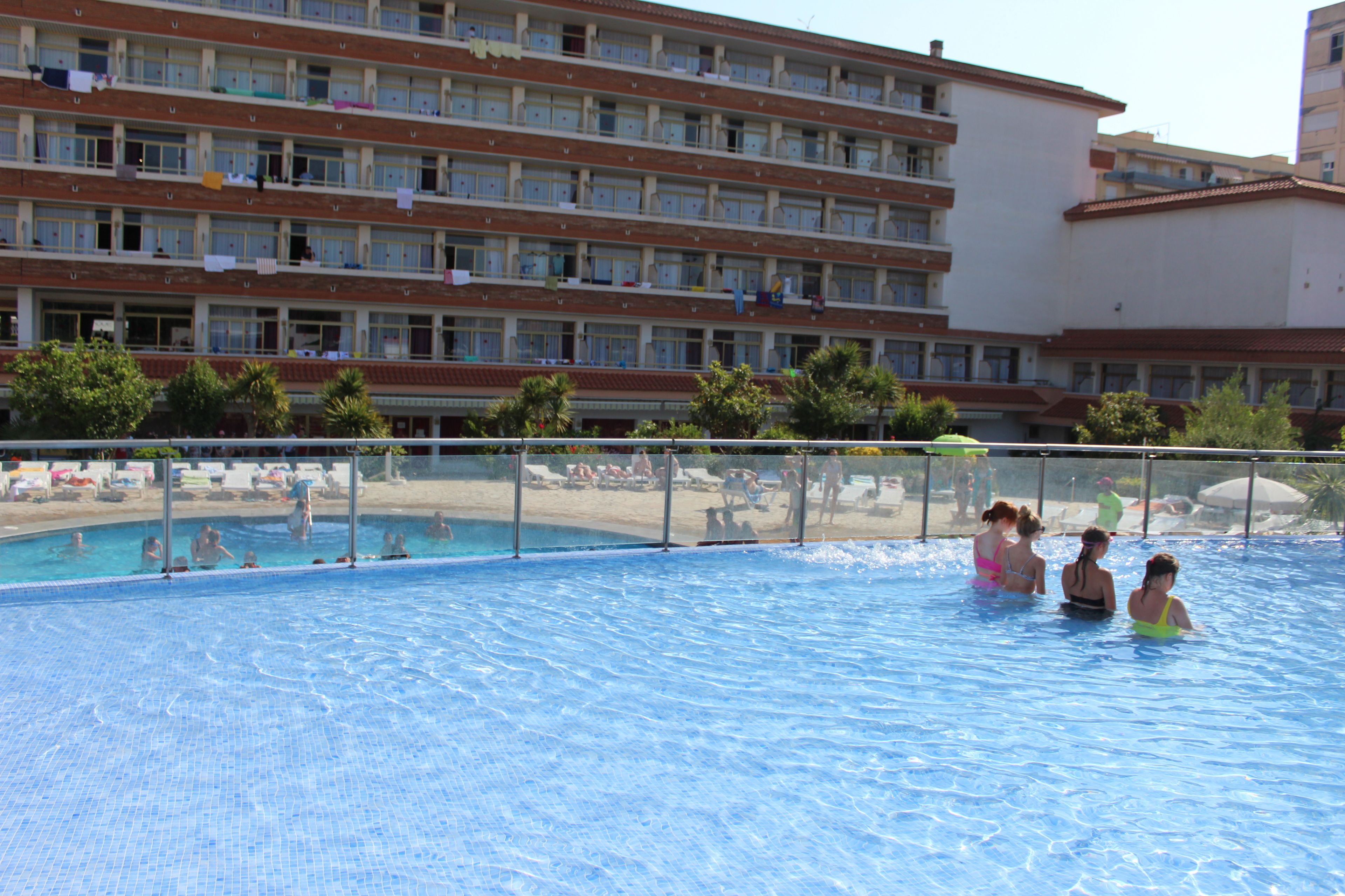 Una piscina con gente en el agua, rodeada por una cerca. Al fondo, un edificio de varios pisos con balcones y ropa tendida.