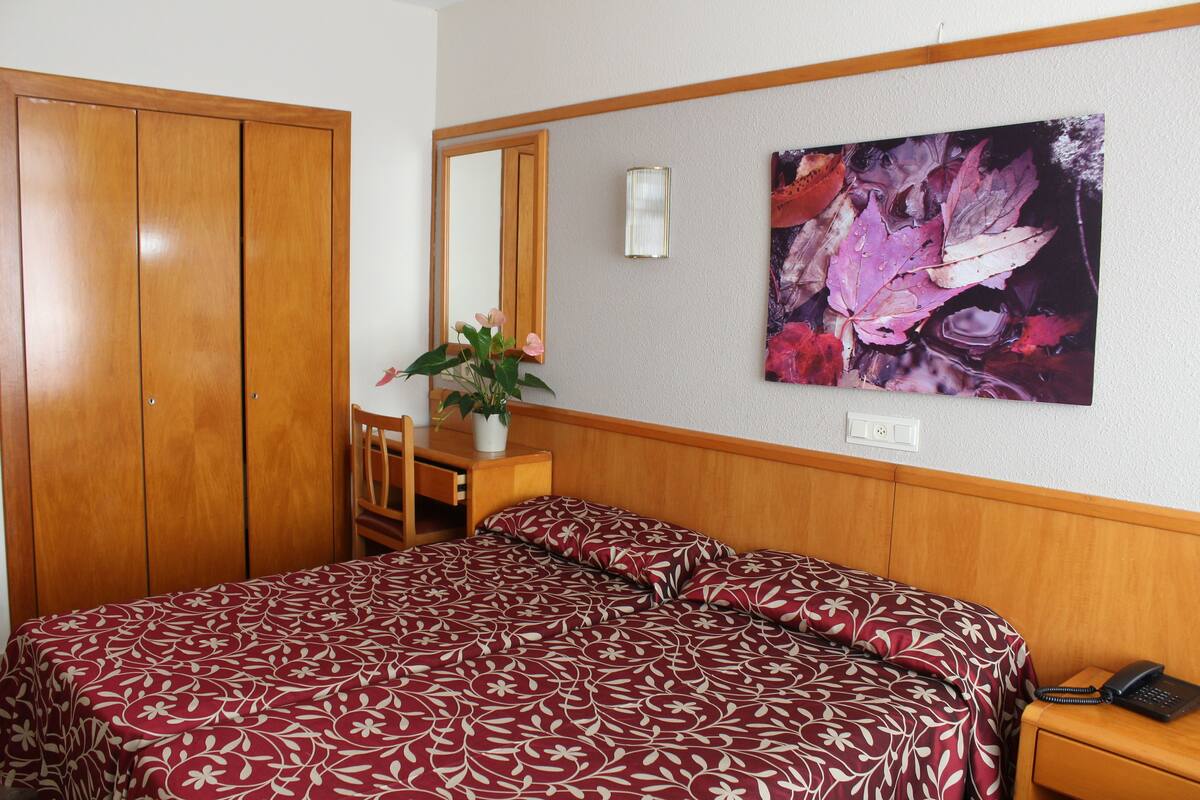 A bedroom with a wooden wardrobe, a desk with a chair, a bed with a patterned bedspread, a wall-mounted telephone, and a framed artwork.