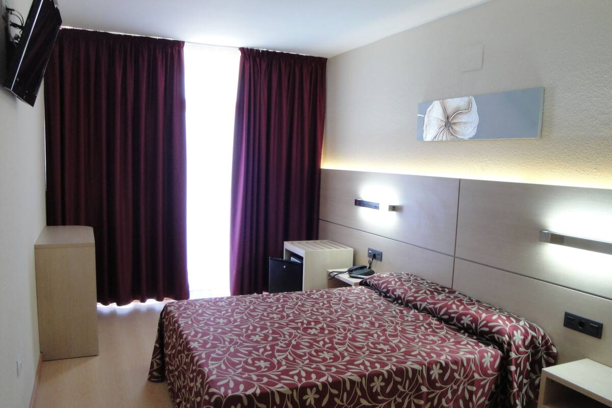 A hotel room with a bed, bedside tables, a desk, a lamp, a TV, and a window with curtains.