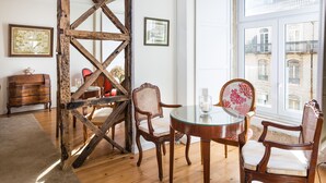 A dining area with a wooden trestle, two chairs with rattan backs, a round glass table, and a view of buildings through a large window. - York House (Lisbon)