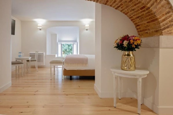 Una habitación de hotel con piso de madera, una cama con cabecera blanca, una mesita con un florero y una ventana con vista a follaje.