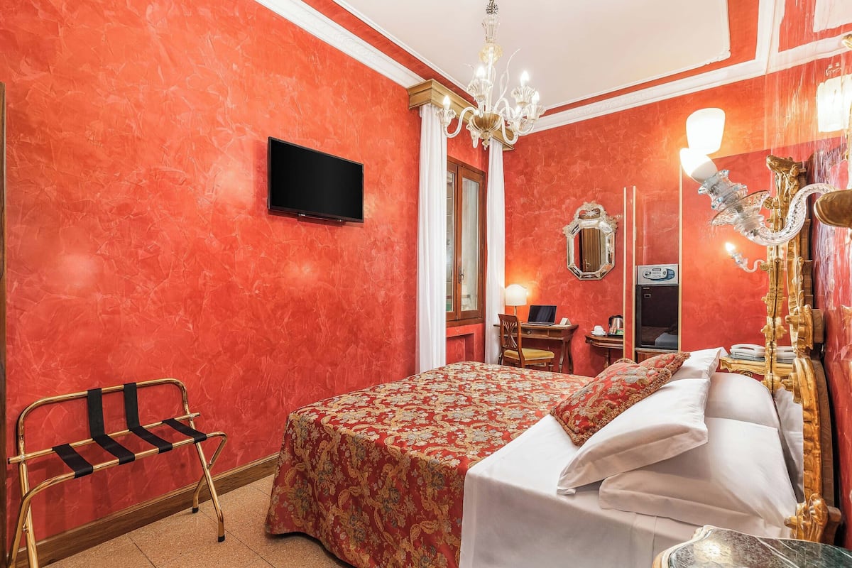 A hotel room with a red wall, a bed with white bedding, a chandelier, a desk with a computer, and a mirror.
