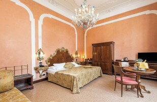 Quarto de hotel com uma cama grande, um guarda-roupa de madeira, uma escrivaninha com cadeira e um candelabro.