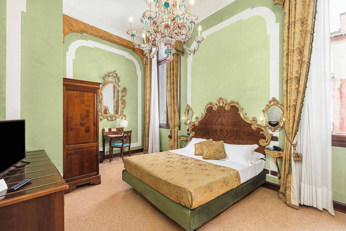 A hotel room with a large bed, a wooden desk with a television, a chandelier, and ornate decor.