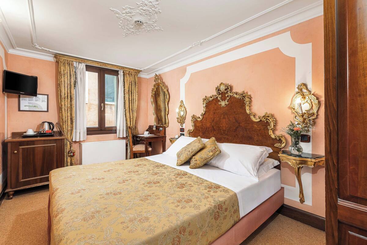 A hotel room with a large bed, a wooden nightstand with a lamp, a desk with a chair, and a television on the wall.