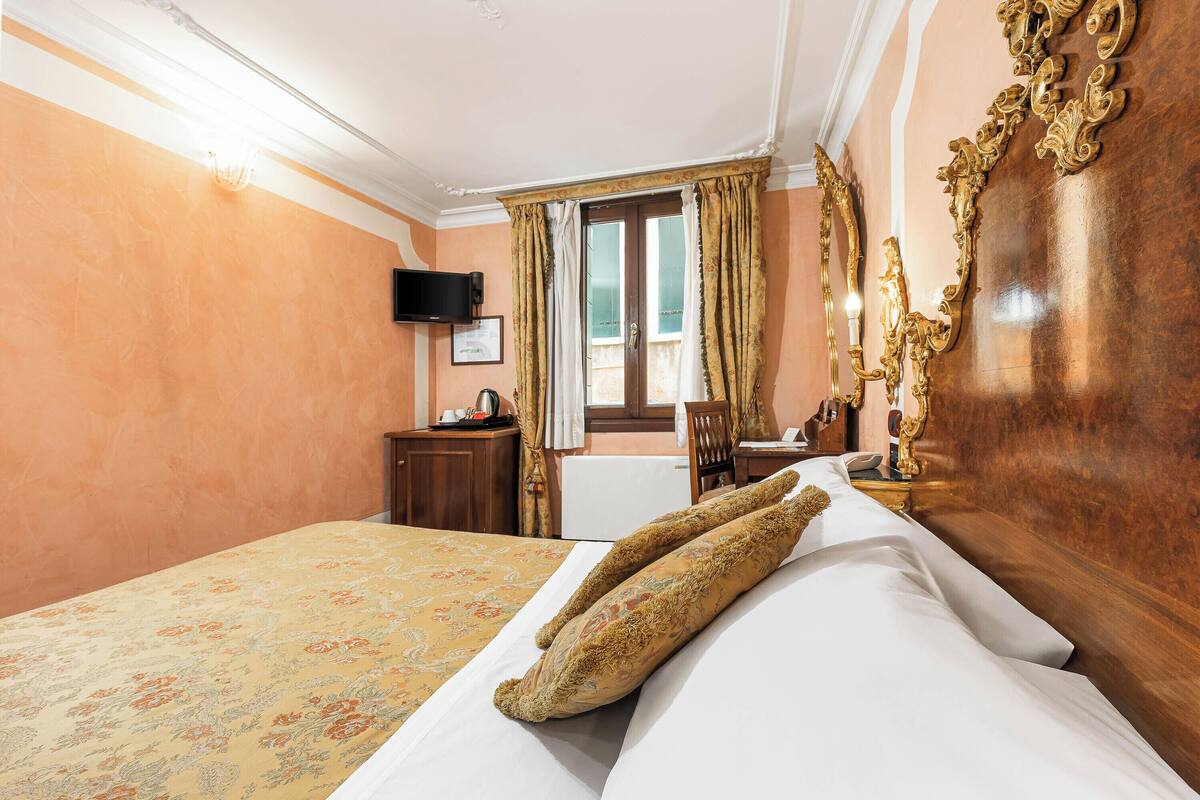 A hotel room with a bed, a wooden headboard, a desk, a chair, a television, and a window with curtains.