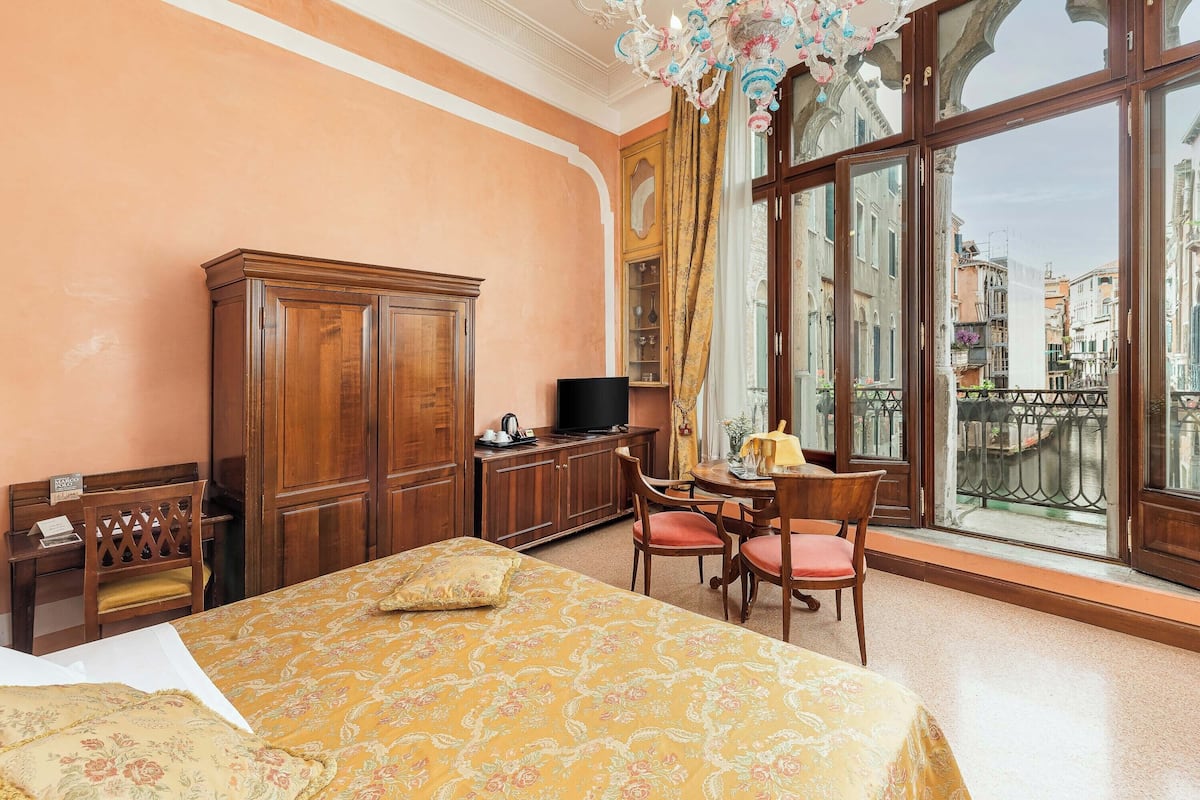 A hotel room with a large bed, wooden furniture, a television, and a view of the city through large windows.