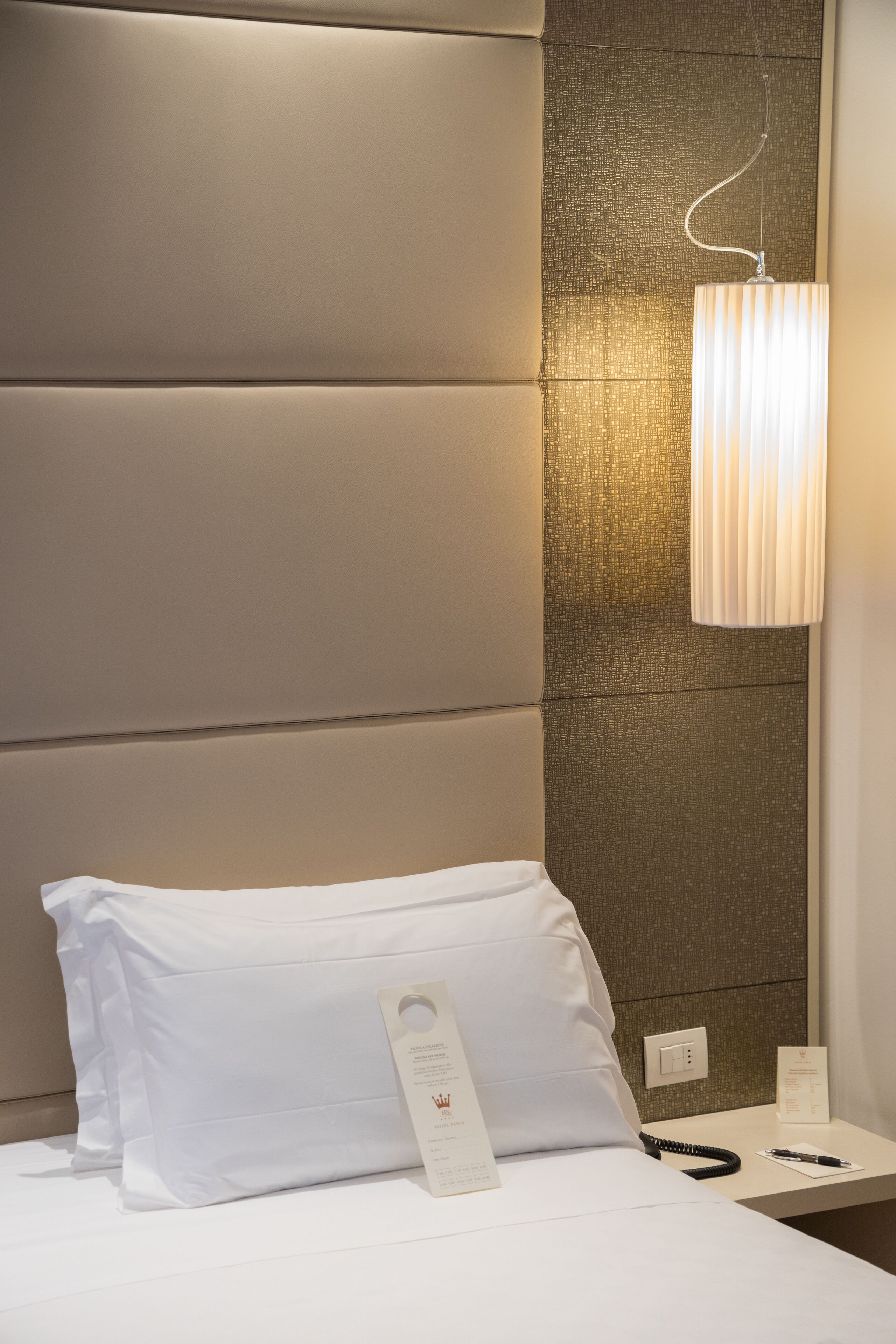 Et hotelværelse med en stor polstret sengegavl, et natbord med en lampe, en pude, en telefon og et kort.