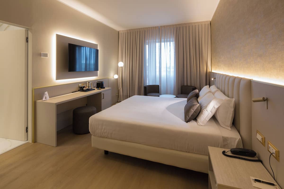 A modern hotel room with a large bed, a desk with a flat-screen TV, a chair, and a bedside table with a phone.