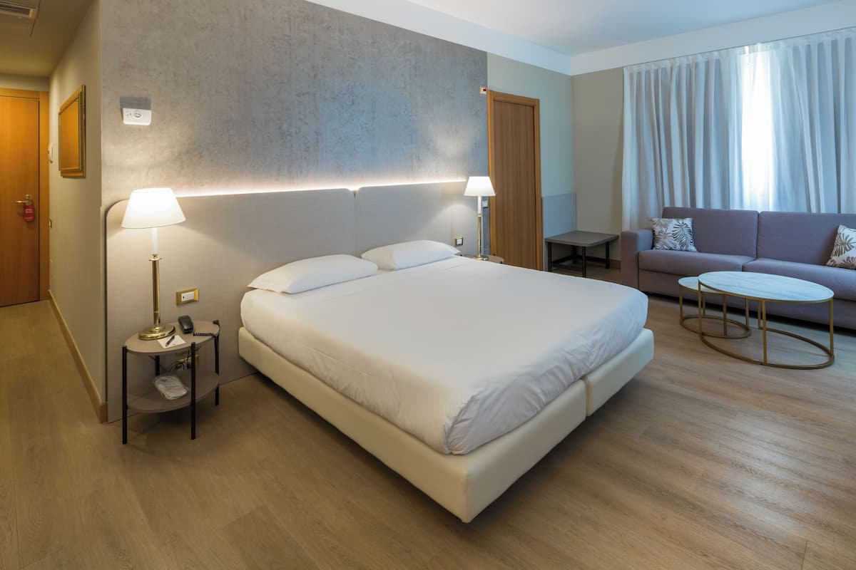 A hotel room with a large bed, bedside lamps, a sofa, a small table, and a view of the outdoors through curtains.
