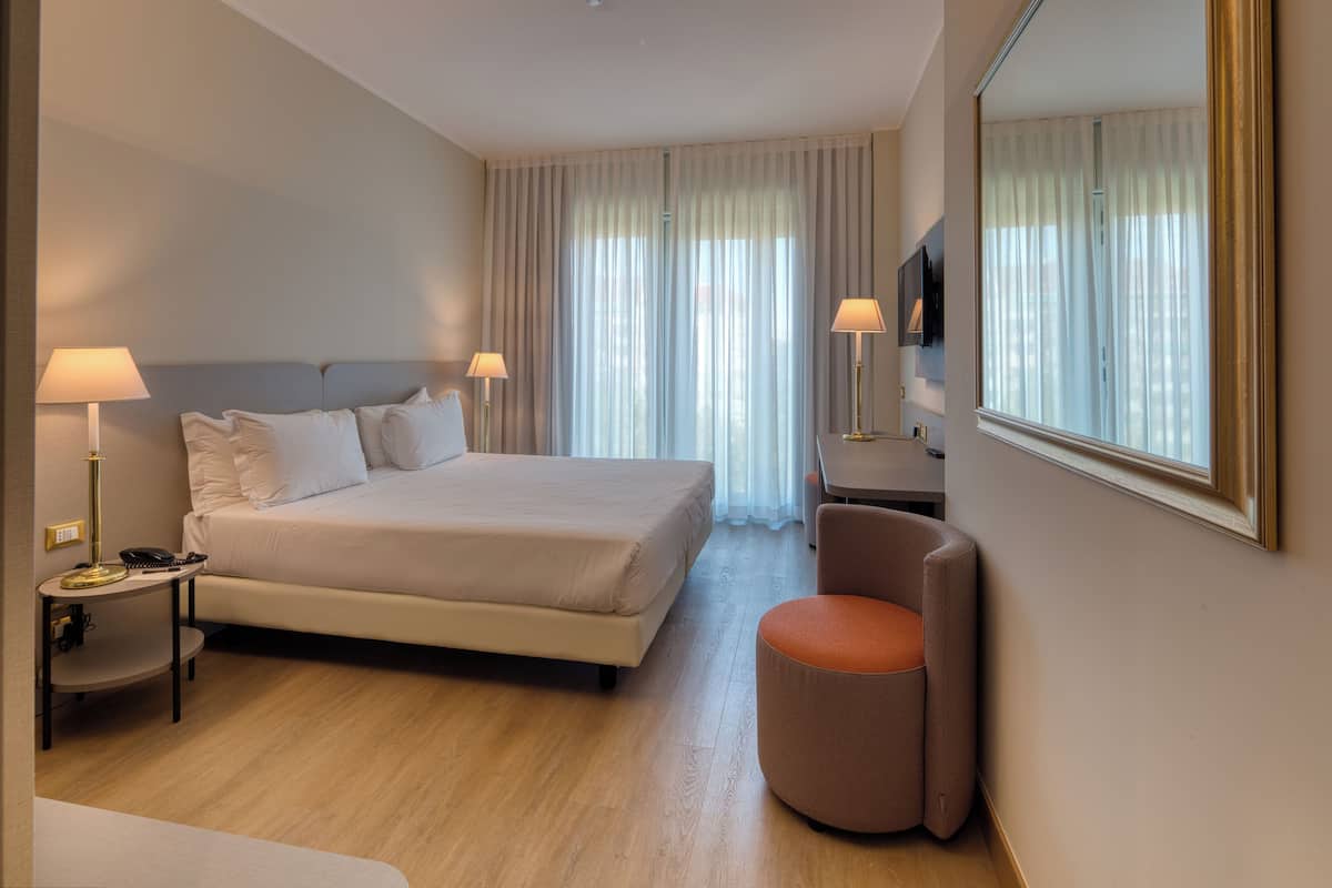 A hotel room with a large bed, two bedside tables with lamps, a chair, a TV, and a large window with curtains.
