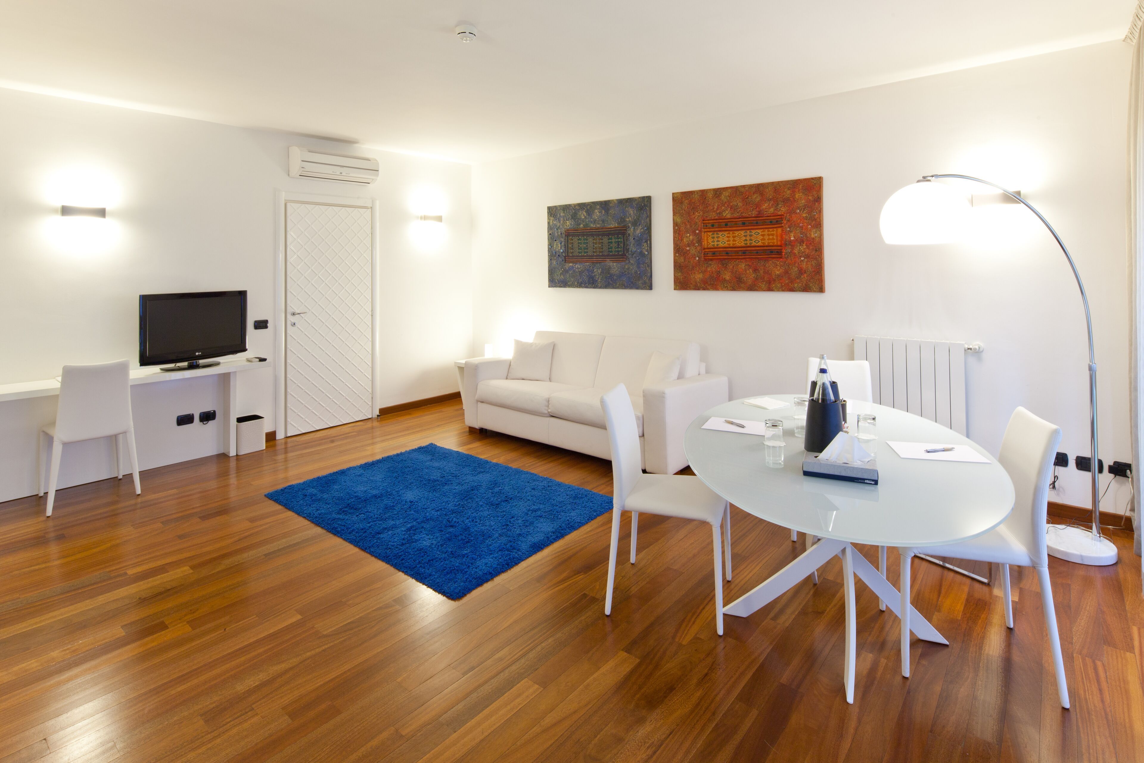 Ein modernes Wohnzimmer mit einem weißen Sofa, einem Esstisch mit Stühlen, einem blauen Teppich und einem Flachbildfernseher.