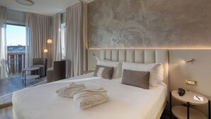 Une chambre d’hôtel moderne dotée d’un grand lit, d’un coin salon avec un fauteuil et une petite table, d’une table ronde avec une lampe, et offrant une vue sur la ville grâce à de grandes fenêtres.