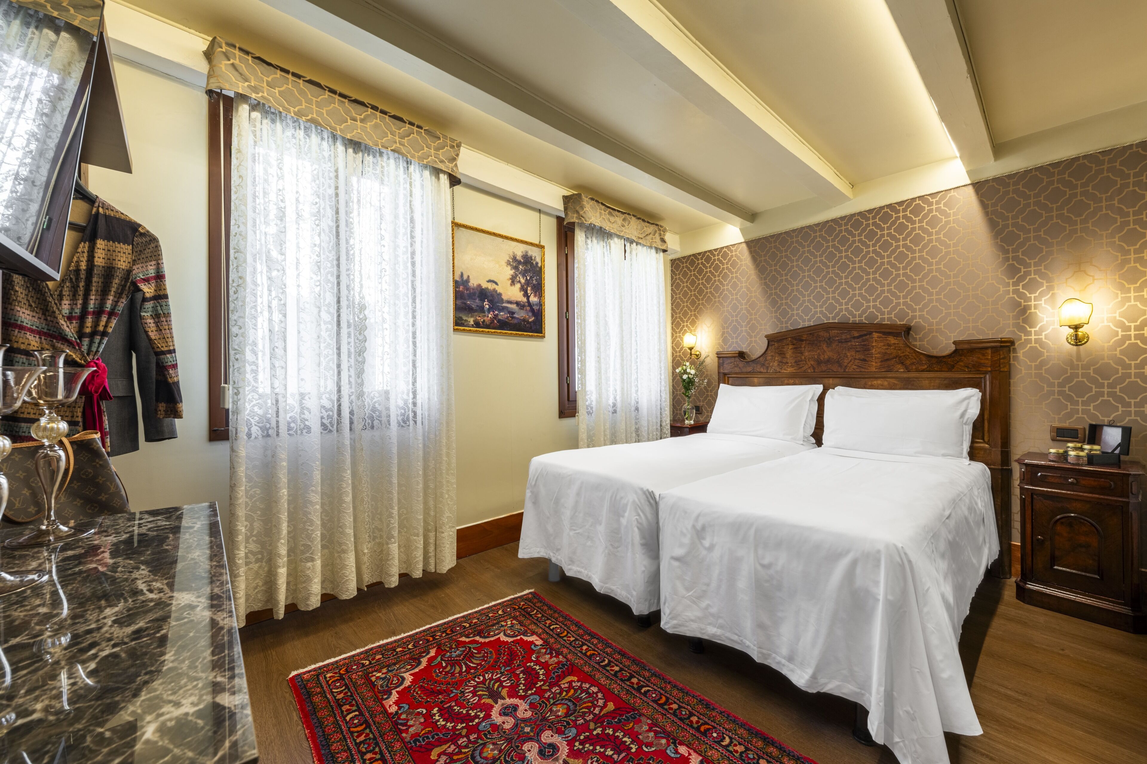 Quarto de hotel com cama de casal, mesa de mármore, tapete com padrão vermelho e mesa de cabeceira de madeira com abajur.