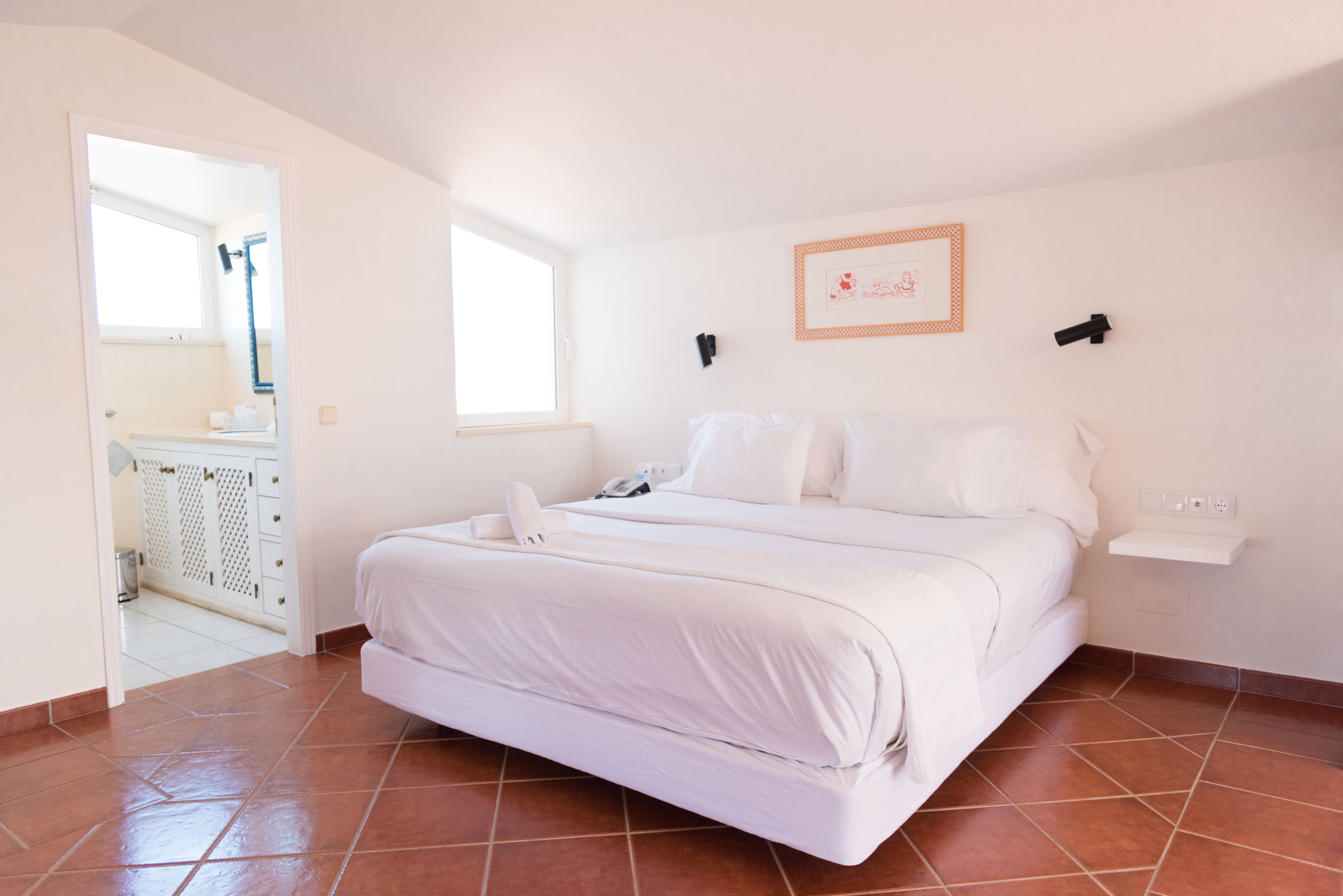 A bedroom with a bed, a window, a wall-mounted shelf, and a door leading to a bathroom.