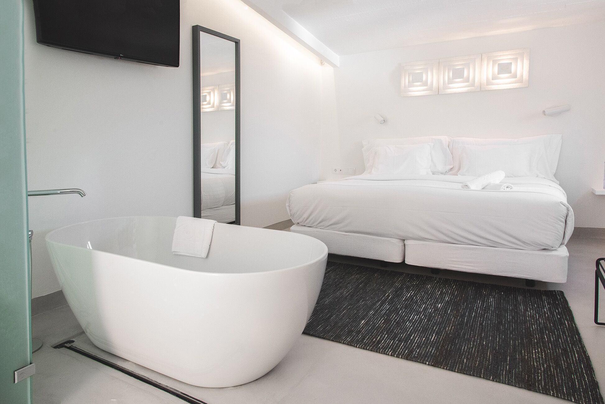 Un dormitorio moderno con una bañera grande, cama con ropa blanca, espejo y lámpara fijada a la pared.