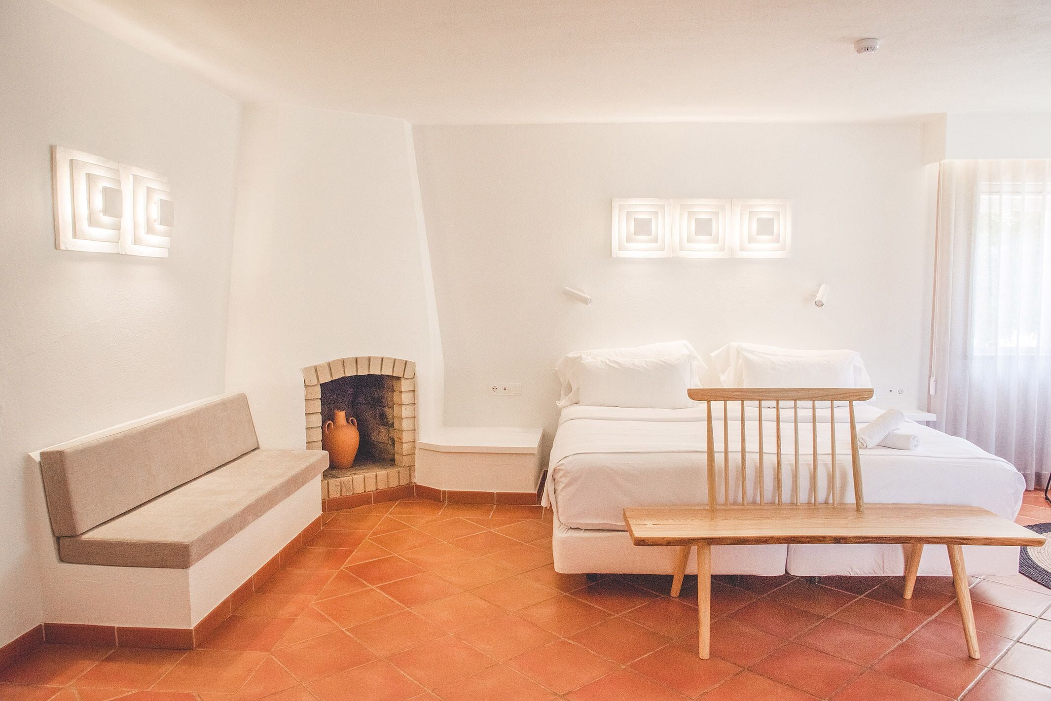 A bedroom with a bed, a bench, a small table, and a fireplace.