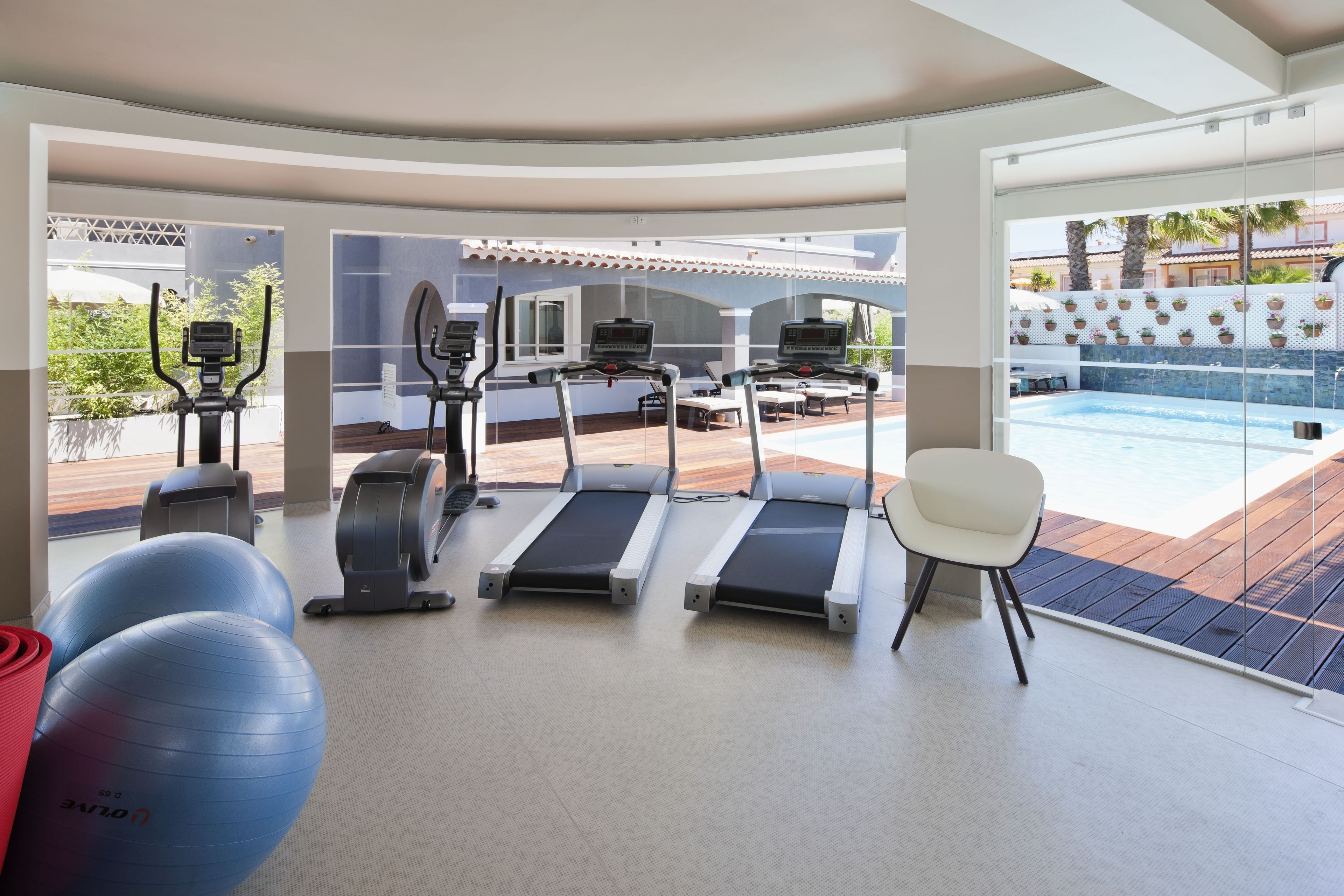 Un gimnasio con cintas de correr, pelotas de ejercicio y una silla, con vistas a una piscina y zona exterior.