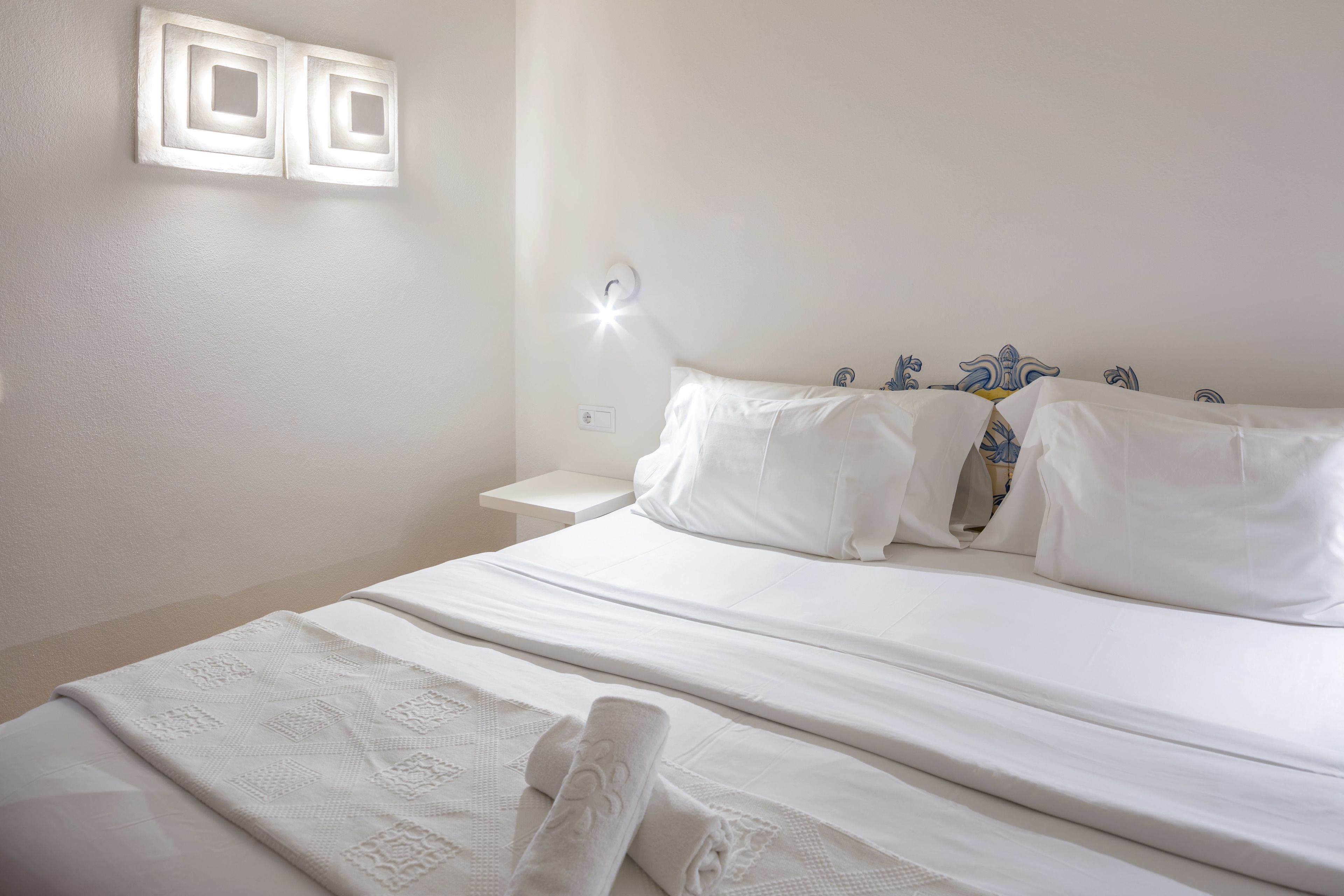 A neatly made bed with white linens and a decorative headboard, a bedside table with a lamp, and a wall-mounted light fixture.