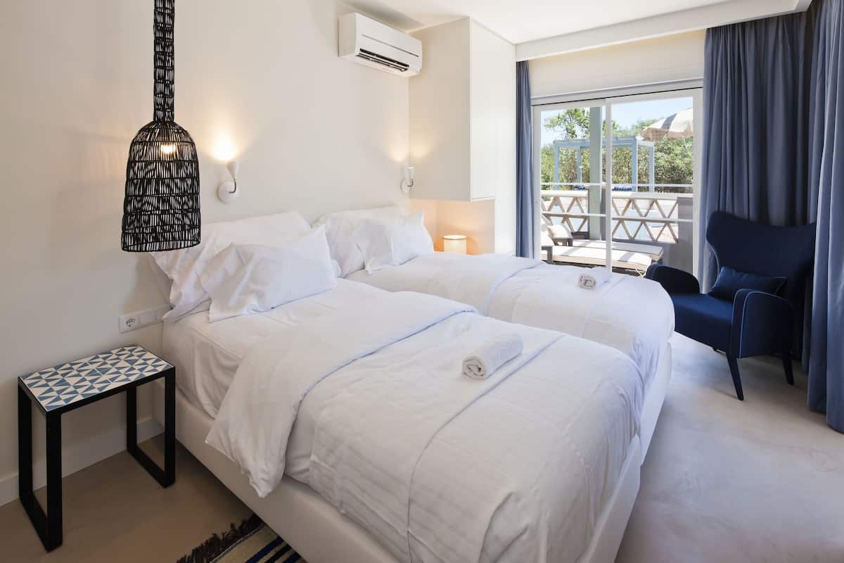 A hotel room with two beds, a small table, a chair, and a balcony with a view.