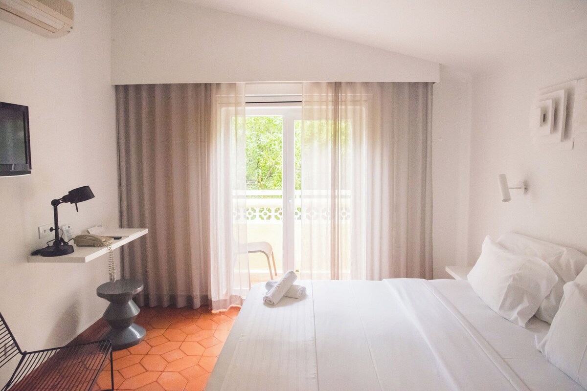 A hotel room with a bed, a desk with a lamp, a chair, and a view of the outdoors through a window with curtains.